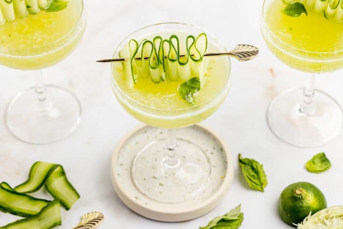 A cucumber Basil gimlet with a ribboned cucumber garnish and two other drinks in the background.